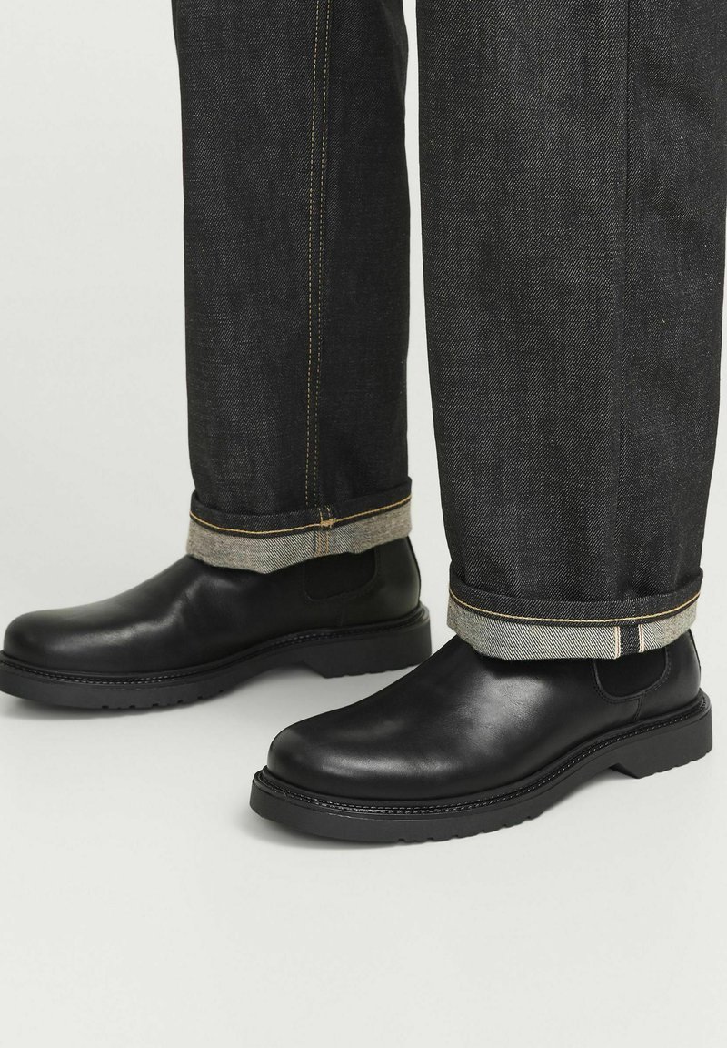 Botas de cuero negras hasta el tobillo con puntera redonda y suela de goma, combinadas con unos vaqueros de mezclilla oscuros que tienen un dobladillo enrollado y costuras en contraste.