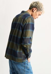 Camicia a quadri verde e blu navy con maniche lunghe, colletto a bottoni e vestibilità rilassata. Orlo laterale spaccato e texture morbida. Indossata con jeans blu.