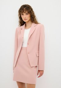 Blazer rosa y falda coordinada en tejido suave. El blazer presenta dos botones y solapas de muesca. Blusa blanca sencilla con escote de cuentas.