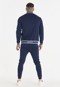 Mężczyzna z tatuażami na szyi i ręce, ubrany w granatowy dres SikSilk i białe tenisówki, stojący tyłem do przodu na białym tle.