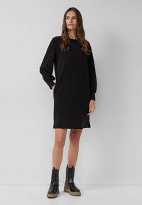 Schwarzes Longsleeve-Kleid mit geradem Schnitt, weicher Textur und seitlichen Taschen; kombiniert mit schwarzen Ankle Boots mit dicken Sohlen und elastischen Seiten.