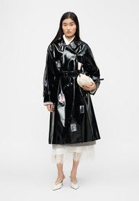 Γυναίκα που φοράει λαμπερό μαύρο καμπαρντίνα με ζώνη πάνω από λευκή δαντελένια φούστα, κρατώντας ένα μικρό λευκό τσαντάκι, στέκεται μπροστά σε απλό φόντο.