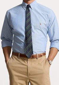 Camicia azzurra chiara con maniche lunghe, cravatta a righe blu navy e verdi, pantaloni chinos beige e cintura in pelle marrone. Orologio al polso.