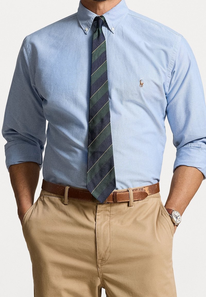 Camicia azzurra chiara con maniche lunghe, cravatta a righe blu navy e verdi, pantaloni chinos beige e cintura in pelle marrone. Orologio al polso.