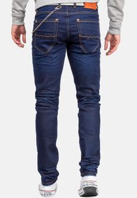 Jeans in denim blu scuro con design aderente, dotati di tasche posteriori e dettaglio in catena metallica. Abbinati a sneakers bianche.