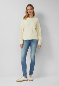 Roomkleurige gebreide trui met een ronde halslijn en geribbelde manchetten, gecombineerd met lichtblauwe slim-fit jeans en witte sneakers met beige accenten.