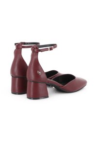 Zapatos de tacón bajo de cuero burdeos con correas en el tobillo y hebilla plateada, punta afilada y un pequeño logo metálico en el tacón.