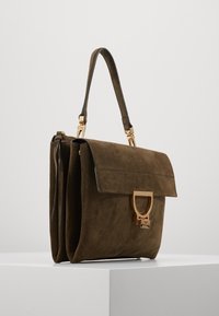 Brązowa zamszowa torebka z górnym uchwytem, złotym zapięciem i geometrycznym, prostokątnym kształtem na białym piedestale.