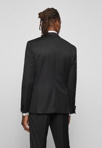 Giacca da uomo nera su misura realizzata in tessuto liscio, con spalle strutturate e doppie aperture sul retro. Include polsini con bottone e un design pulito sulla schiena.
