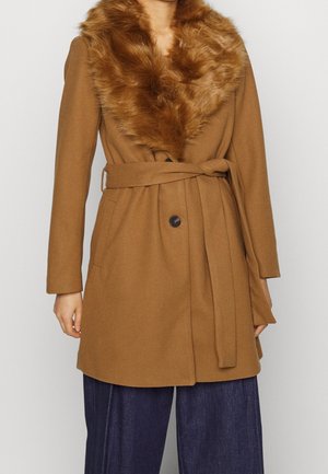 Manteau en laine couleur camel avec ceinture et grand col en fausse fourrure, porté sur un pantalon large foncé, mains détendues sur les côtés.