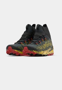 Pantofi de alergare pe teren înalți, cu un design în culori negru și galben cu stropi, tălpi roșii și mânecă integrată pentru gleznă, tot neagră.