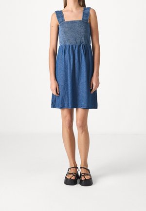 Kvinde iført knælængde blå denimkjole med smocket overdel og brede stropper, kombineret med sorte plateau-sandaler mod hvid baggrund.