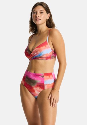 Kobieta stoi w kolorowym, wysoko taliowanym bikini z różowymi, czerwonymi i niebieskimi abstrakcyjnymi wzorami, pozując na jasnym, jednolitym tle.