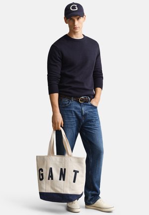Junger Mann in einem marineblauen Pullover, blauen Jeans und Mütze, der eine beige Tragetasche mit dem Schriftzug "GANT" hält und vor einem weißen Hintergrund steht.