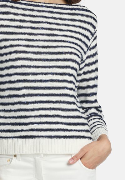 Femme portant un pull en maille à rayures horizontales blanches et bleu marine avec des paillettes, assorti à un pantalon blanc, une main dans la poche.