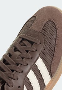 Sneaker marrone con tomaia in mesh e pelle, superficie testurizzata, accento bianco a tre strisce e suola in gomma. Lacci dello stesso colore marrone.