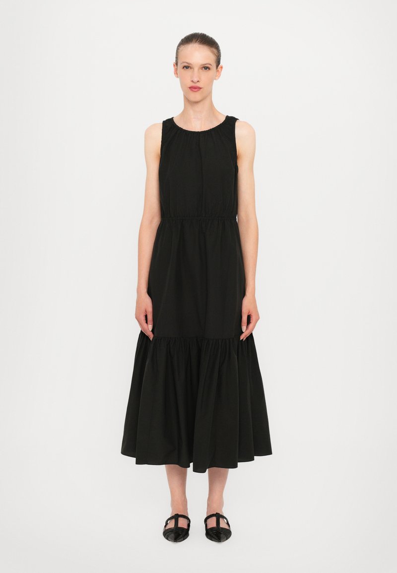 Black sleeveless dress made of cotton with a gathered waist, flowing skirt, and a textured bodice. Worn with black woven shoes.