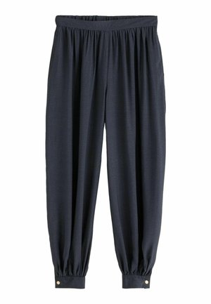 Persoon die losse zwarte broek en een donker crop top draagt, staat blootsvoets op een zandstrand vlak bij het water.