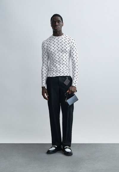 Man wearing a white long-sleeve top with black crescent patterns, black textured trousers, black-and-white shoes, holding a small black handbag.