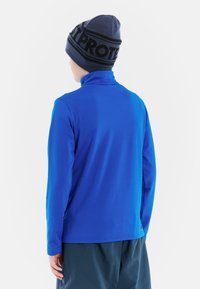 Camicia blu a maniche lunghe con colletto alto, realizzata in tessuto morbido elastico. Indossata con un berretto lavorato a maglia con un motivo nero e blu.