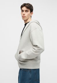 Jauns vīrietis, valkājot gaiši pelēku džemperi ar rāvējslēdzēju un zilas džinsa bikses, pozē ar rokām džempera kabatās uz vienkrāsaina fona.