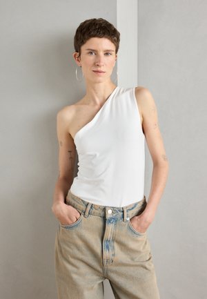 Asymmetrical white bodysuit made of stretchy fabric, paired with light wash denim pants. Model stands with hands in pockets against a neutral background.
