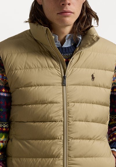 Beige puffer vest with vertical quilted pattern, zip closure, high collar, embroidered logo on chest, layered over a colorful, patterned sweater.
