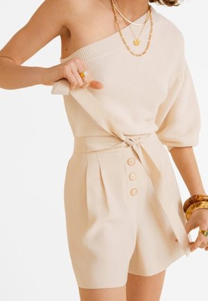 Frau richtet den gebundenen Gürtel an beige, hoch taillierten Shorts mit drei Knöpfen, trägt ein einseitiges Stricktop und goldenen Schmuck.