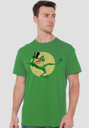Mann trägt grünes T-Shirt mit tanzendem Cartoon-Frosch im Zylinder vor gelbem Kreis-Hintergrund.