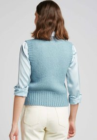 Un gilet tricoté bleu clair avec une finition côtelée, en fil texturé, porté par-dessus un chemisier satiné bleu clair. Un pantalon blanc avec des poches arrière.