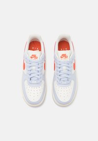 Fehér és világoskék sportcipők piros részletekkel, perforált orral, fehér fűzővel és textúrázott hálós béléssel. A márka hangsúlyosan megjelenik.