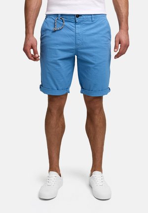 Mannelijk model met blauwe opgerolde short met trekkoord, witte sneakers en een wit T-shirt, staand tegen een effen achtergrond.