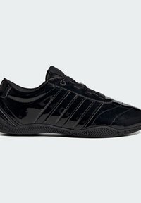 Baskets noires brillantes avec laçage à l'avant, semelle texturée et trois bandes parallèles sur le côté sur un fond clair uni.
