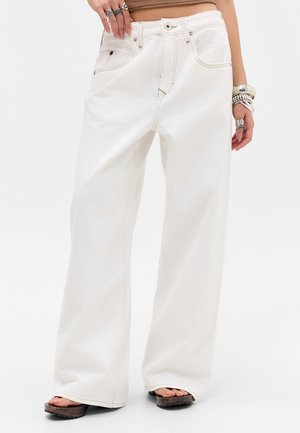 Valged laia säärega teksad, mida kantakse platvormsandaalidega, käevõrude ja mitmete sõrmustega kätes, ühevärvilise tausta ees.