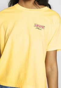 Nainen pukeutuneena löysään keltaiseen Levi'sin Kalifornia T-paitaan, jossa on vaaleanpunainen logo, näkyy osittain kaulasta vyötäröön.