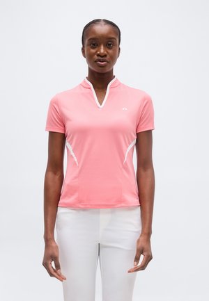 Donna che indossa una maglietta sportiva rosa a maniche corte con scollo a V e accenti bianchi, pantaloni bianchi, in piedi davanti a uno sfondo semplice.