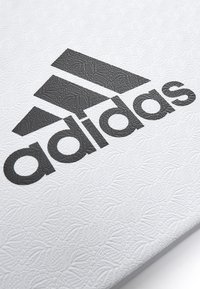 Adidas-Logo auf einer strukturierten weißen Oberfläche mit einem detaillierten Blattmuster, versehen mit fett gedruckten schwarzen Buchstaben und geometrischen Akzenten.