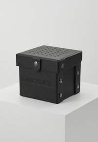 Boîte en cuir noir avec un couvercle perforé, dotée d'un fermoir et d'accents métalliques. Logo "DIESEL" en relief à l'avant. Forme carrée.