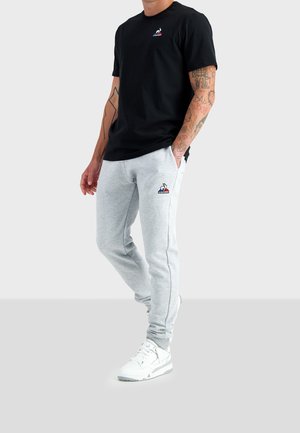 Homme portant un t-shirt noir et un pantalon de jogging gris clair avec des logos, des baskets blanches et des tatouages visibles sur les bras, debout les mains dans les poches.