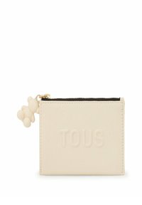 Cartera de cuero color crema con el logo "TOUS" en relieve. Presenta una cremallera negra y un charm de oso de peluche de color claro adjunto.