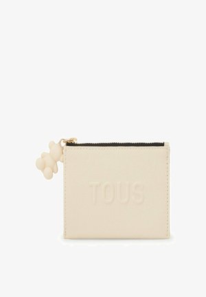 Cartera de cuero color crema con el logo "TOUS" en relieve. Presenta una cremallera negra y un charm de oso de peluche de color claro adjunto.