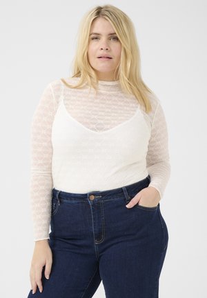Haut à manches longues en dentelle blanche avec motif géométrique et col montant, porté sur un débardeur à col dégagé, associé à un jean taille haute bleu marine.