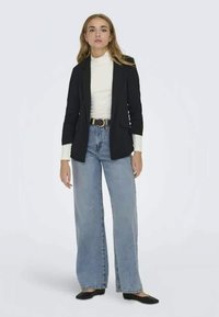 Μαύρο σακάκι με λεπτομέρειες στις τσέπες, επενδεδυμένο πάνω από ένα λευκό πουλόβερ και συνδυασμένο με ψηλόμεσα, ανοιχτόχρωμα φαρδιά τζιν και μαύρες μπαλαρίνες.