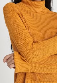 Femme portant un pull en maille jaune moutarde à col roulé, les bras croisés, montrant une texture détaillée et des poignets côtelés.