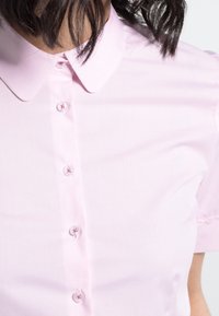 Femme portant une chemise rose clair à manches courtes, boutonnée, avec col arrondi et coupe ample.