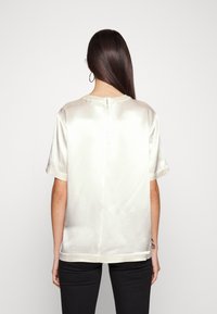 Top corto in raso a maniche corte bianco sporco con colletto rotondo, chiusura con zip sul retro e texture liscia; abbinato a pantaloni neri aderenti.