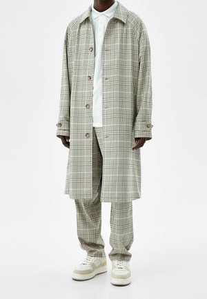 Homme portant un manteau long à carreaux beige et un pantalon assorti avec un polo blanc et des baskets blanches sur fond blanc.