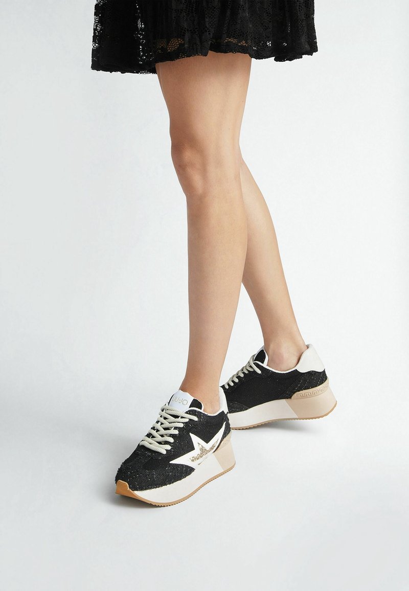 Jambes portant une jupe en dentelle noire et des baskets à plateforme noires avec des détails en forme d'étoile blanche et des semelles beiges sur fond blanc.