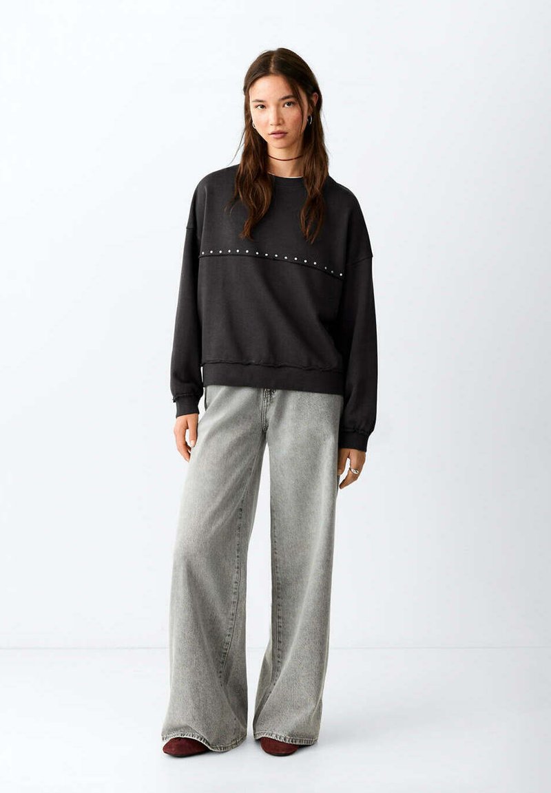 Jeune femme aux longs cheveux portant un sweat-shirt noir clouté et un jean gris à jambes larges, debout devant un fond blanc uni.