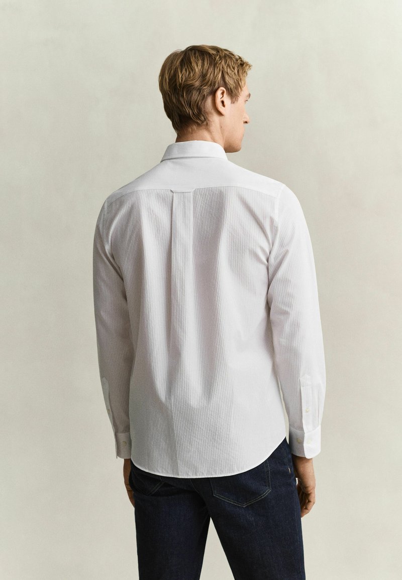 Man with short light hair wearing a white long-sleeve shirt and dark jeans, facing away against a plain neutral background.
